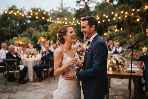 Pareja nupcial brindando riendo con copas de champagne bajo guirnaldas de luces en banquete al aire libre